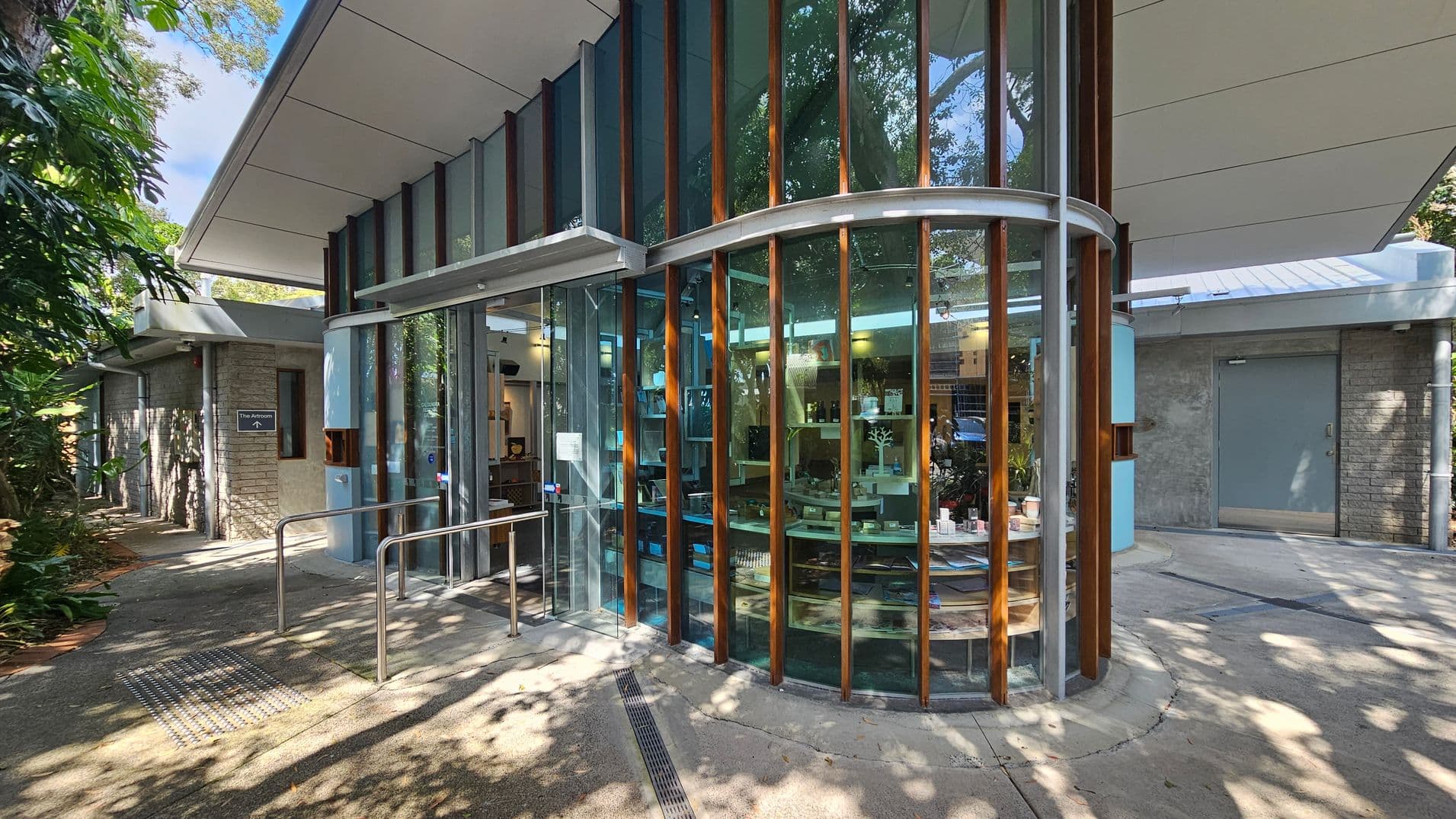 A modern building with a curved glass facade featuring vertical wooden slats. Inside, shelves display various items. The entrance has a metal handrail and a sign reading "The Atrium" on a stone wall to the left. The roof is flat with a wide overhang, and the ground is paved with concrete. Lush greenery is visible on the left side, providing shade and a natural backdrop.