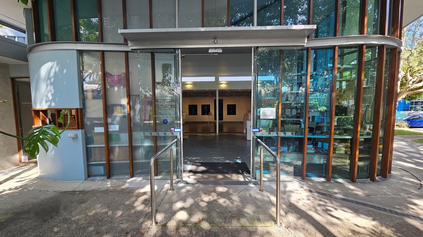 Glass entrance to a building with a curved facade, featuring vertical wooden beams. The doors are open, revealing an interior with a polished floor and artwork on the walls. Metal railings lead up to the entrance. A large plant is visible on the left, and a blue car is parked outside on the right. Shadows of trees are cast on the ground.