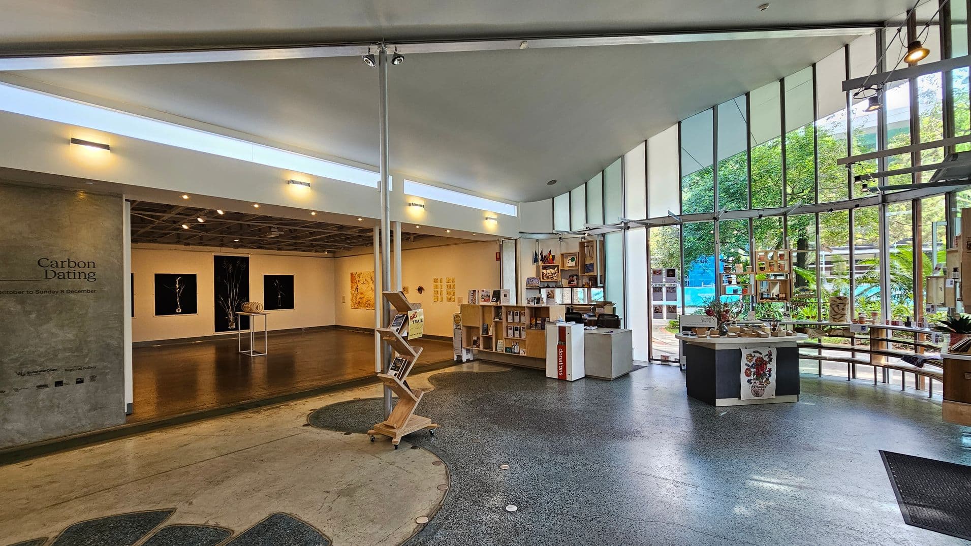 A modern gallery interior with a polished concrete floor. On the left, an exhibition space with dark wooden flooring displays artwork on white walls. The entrance to this area has a sign reading "Carbon Dating". In the centre, a wooden display stand holds brochures. On the right, a reception desk and shelves with books and plants are near large windows, offering a view of greenery outside. The ceiling is high and white, with spotlights illuminating the space.