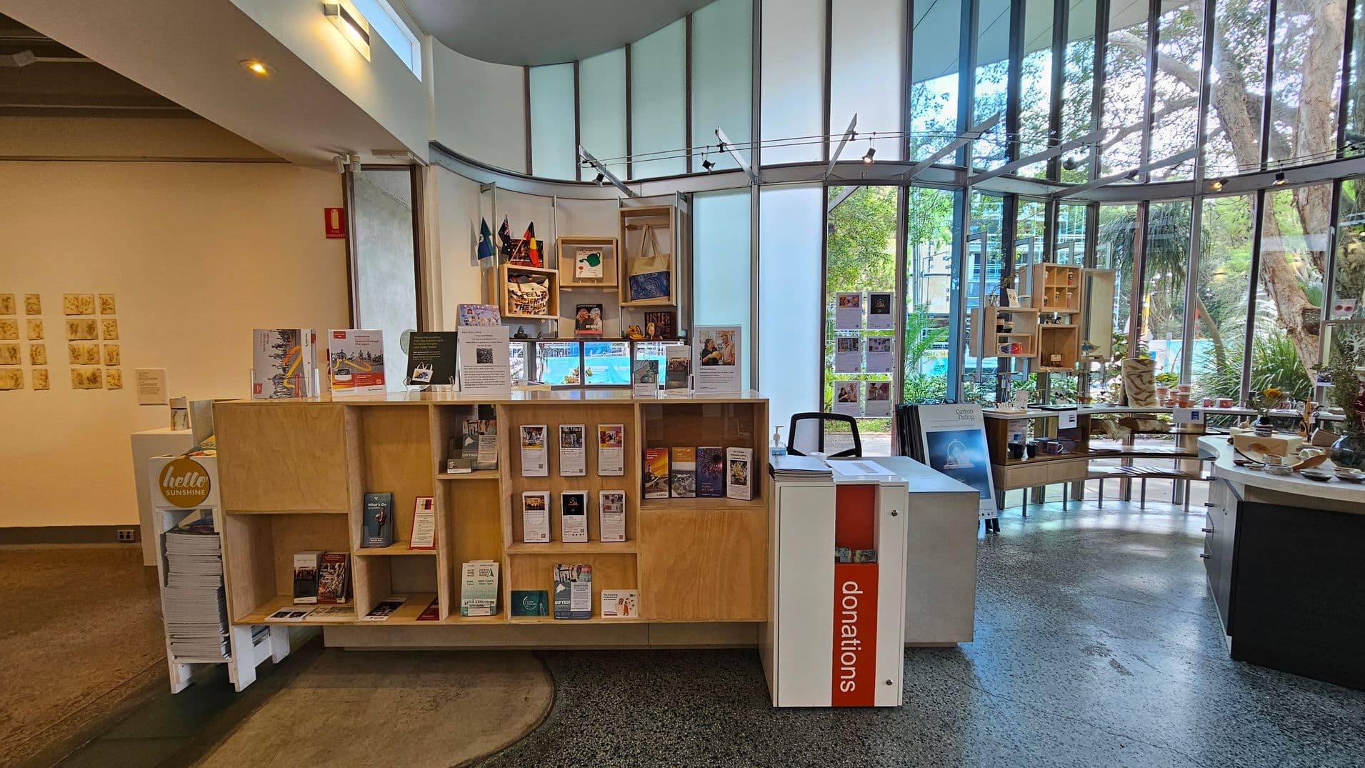 A room with large glass windows in the background, allowing natural light to illuminate the space. In the centre, there is a wooden display shelf filled with brochures and informational materials. To the right, a white donation box is visible. On the left, a wall displays small artworks. The floor is polished concrete, and the ceiling has recessed lighting. Outside, trees and greenery are visible through the windows.