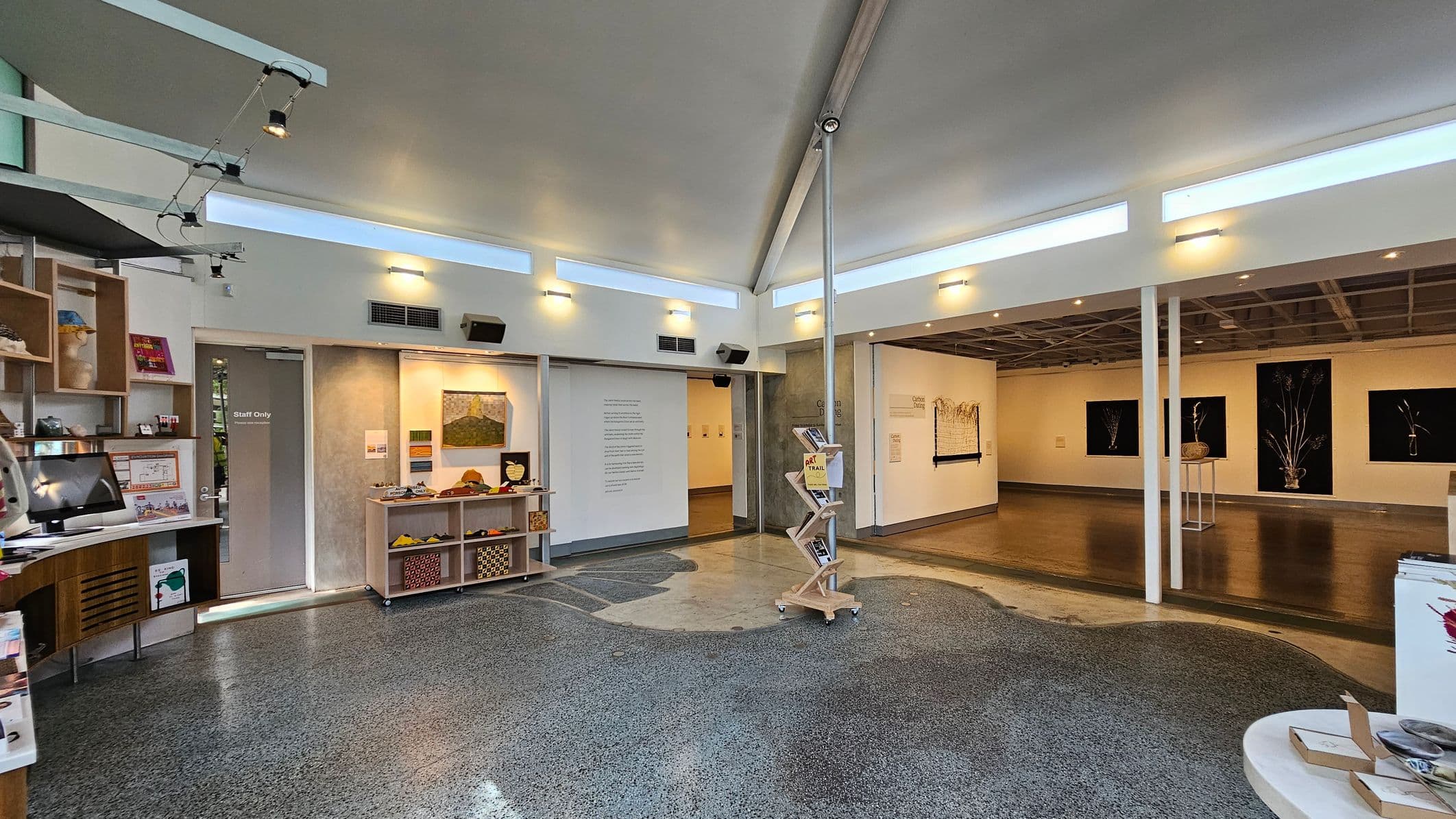 A modern interior space with a polished, speckled floor. On the left, there are shelves with various items and a "Staff Only" door. In the centre, a display stand holds brochures. The right side features an open area with artwork on the walls, including framed pieces with dark backgrounds. The ceiling has exposed beams and track lighting, and the walls are adorned with small, evenly spaced lights.