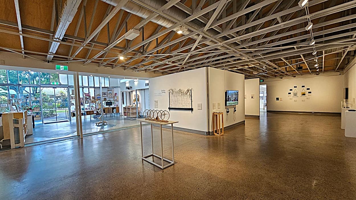 An art gallery with white walls and a polished concrete floor. On the left wall, there's a piece titled "Carbon Dating" with a grid and plant-like elements. The back wall displays three black panels with botanical images and a sculpture on a pedestal. The right wall features a large abstract painting, a small circular artwork, and a grid of smaller images. A wooden shelf with brochures is in the foreground on the right. The ceiling has exposed beams and spotlights.