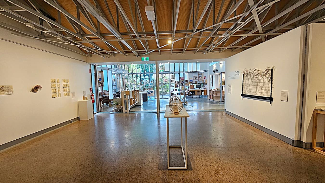An art gallery interior with polished concrete floors and an exposed beam ceiling. On the left, large glass doors and windows reveal an outdoor area with greenery. In the centre, a display table holds circular sculptures. The right wall features a mounted screen and a series of small artworks. The back wall has a collection of framed pieces arranged in a pattern. The space is well-lit with track lighting.