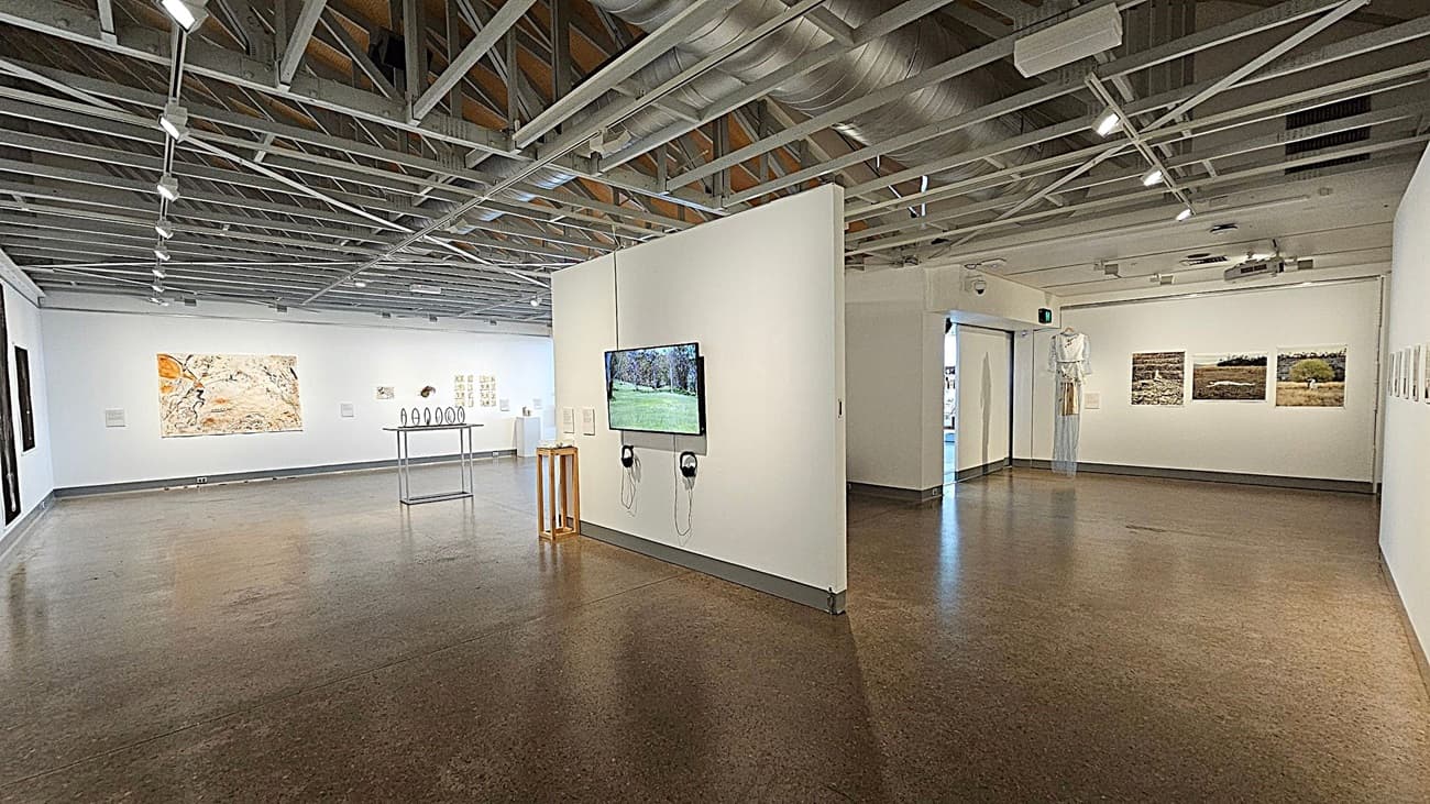 An art gallery interior with a polished concrete floor and an exposed beam ceiling. In the centre, a narrow table displays a spherical sculpture. The left wall features small artworks and a fire extinguisher nearby. The right wall has a woven art piece. Beyond the glass doors, a well-lit area with bookshelves and various items is visible, with greenery outside.
