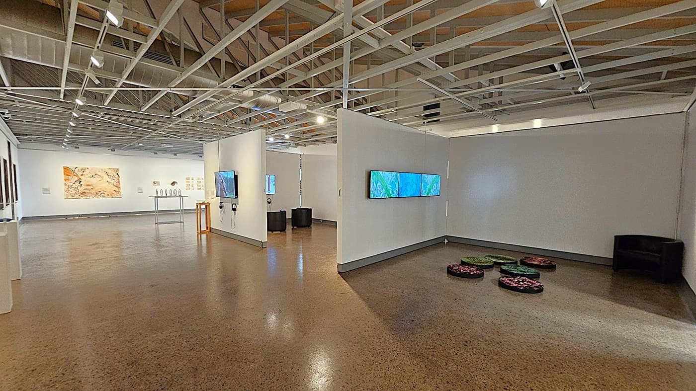 A spacious art gallery with polished concrete floors and an exposed beam ceiling. On the right, a wall displays three digital screens showing blue and green images. Below, circular cushions are arranged on the floor. In the centre, a partition wall holds a screen with headphones. On the left, artworks are mounted on the walls, including a large abstract piece. A table with sculptures is visible in the background. The lighting is bright, highlighting the exhibits.