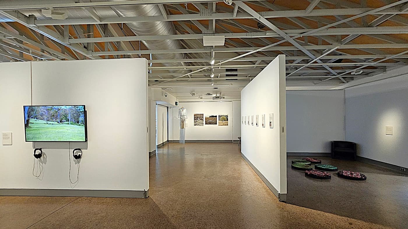 A gallery interior with a polished concrete floor and an exposed ceiling with metal beams and ducts. On the left wall, a screen displays a landscape image, with headphones hanging below. The central area features a corridor with artwork on the walls, leading to a back wall with three framed pictures. On the right, a dark corner contains a black chair and several colourful circular cushions on the floor.
