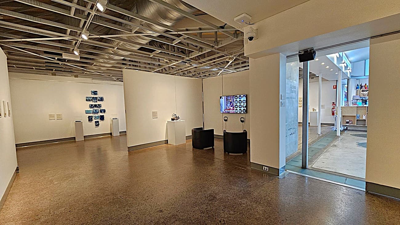 An art gallery interior with white walls and a polished brown floor. On the left, a series of blue artworks are displayed on the wall. In the centre, a small sculpture is placed on a pedestal. Two black chairs face a wall-mounted screen with headphones. The ceiling features exposed ductwork and lighting. On the right, a glass door leads to another room with shelves and a fire extinguisher visible.