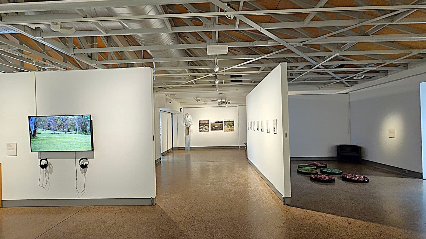 A gallery interior with a polished concrete floor and an exposed ceiling with metal beams and ducts. On the left wall, a screen displays a landscape image, with two sets of headphones hanging below. In the centre, a corridor leads to a back wall featuring three framed artworks. On the right, a dark corner contains a black chair and several round, patterned cushions on the floor. The walls are white, enhancing the display of art pieces.