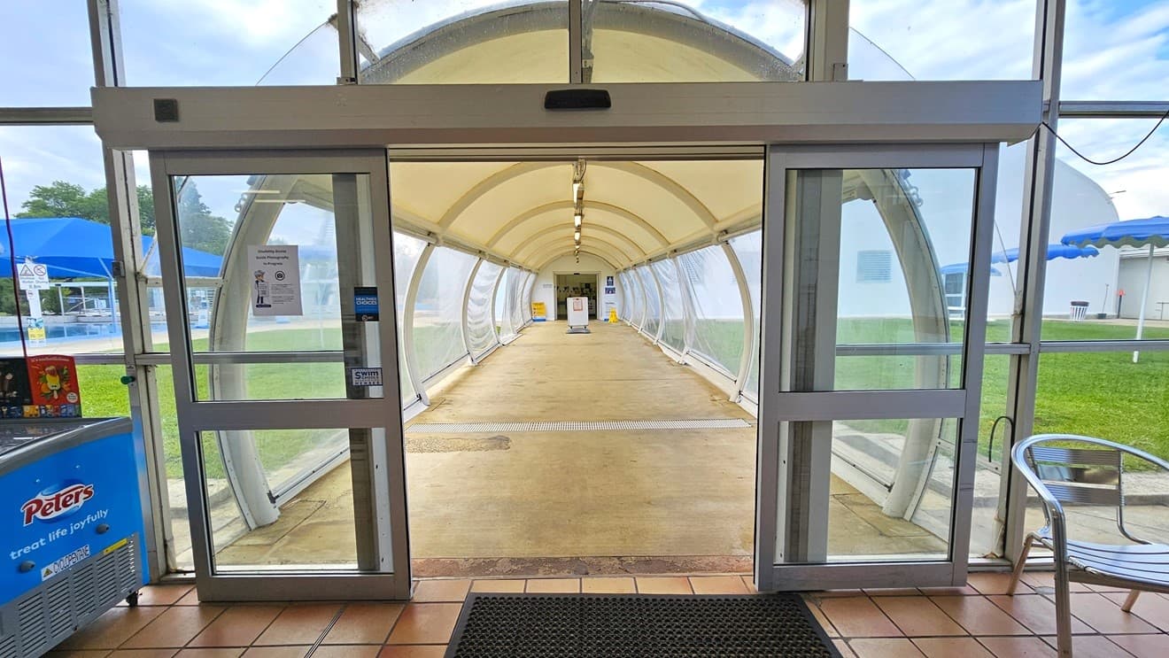 In the middle is an automatic glass door that leads to a tunnel constructed with durable vinyl material, featuring adjustable side panel flaps. An anti-slip mat is placed on the doorsteps and movable silver chairs are visible on the right.  On the left is an ice cream freezer. Both sides of the tunnel are green grass and blue shade cloth. Truncated domes are installed near the door. Further ahead, at the end of the tunnel, banners are visible.
