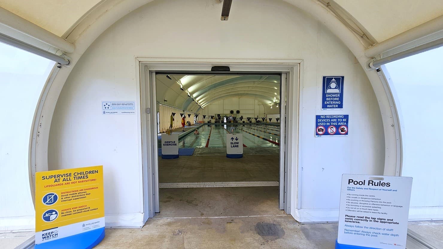 The dome-shaped entrance displays posters and safety reminders. Inside, truncated domes are installed. The lap pool is visible and equipped with floating ropes. Free-standing banners are positioned on the pool deck.