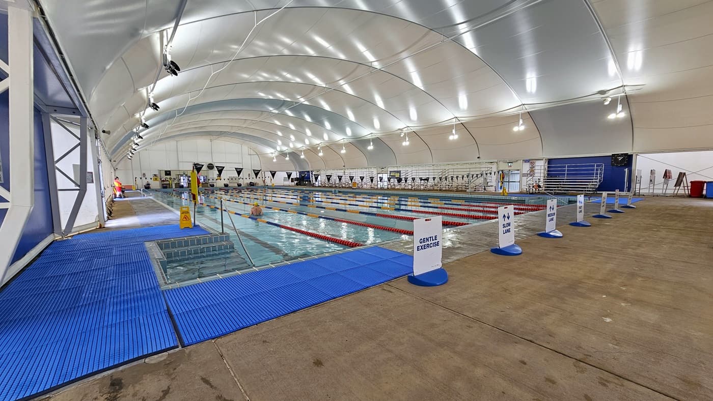 In the middle is the 8-lane lap pool, equipped with floating ropes and stairs with railings. Free-standing banners are positioned on the pool deck. Blue matting is installed in the left corner. Tiered seatings are positioned on the right. Strings of blue flags are hung across the pool. The dome-shaped ceiling features floodlights and LED lights