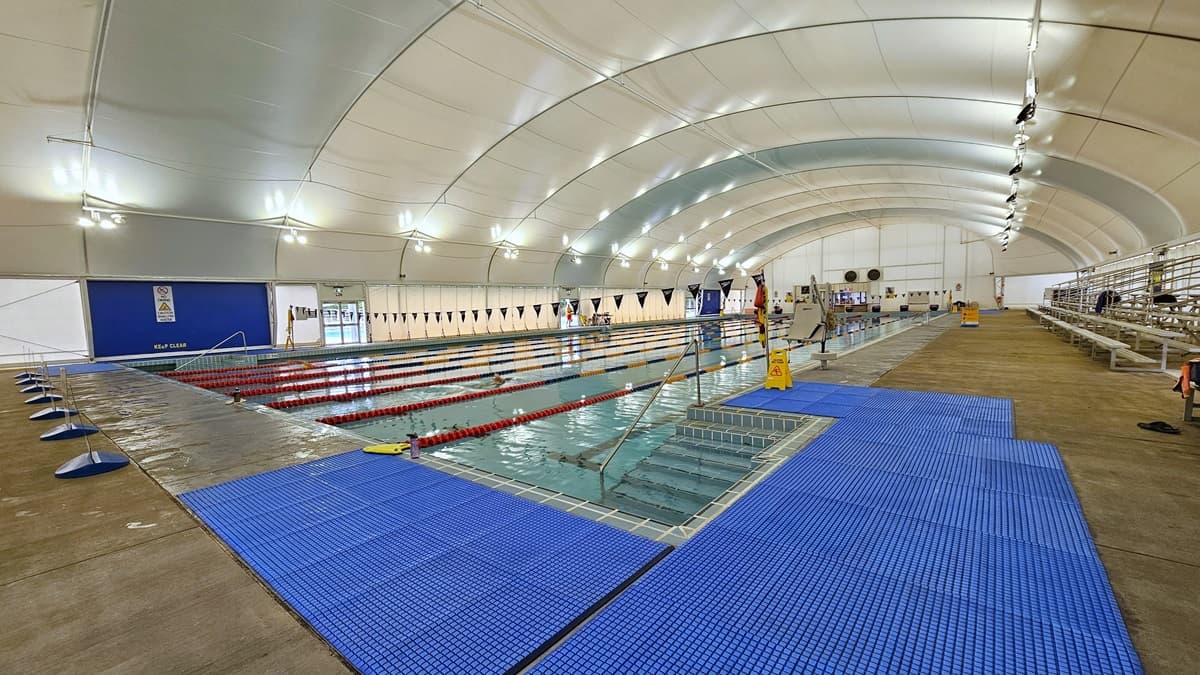 In the center is the 8-lane lap pool, equipped with floating ropes and stairs with railings. Free-standing banners are positioned on the pool deck. Blue matting is installed on both corners. Tiered seating is on the right. Strings of blue flags are hung across the pool. Safety reminder banners are visible on the pool deck area to the right. The dome-shaped ceiling features floodlights and LED lights.