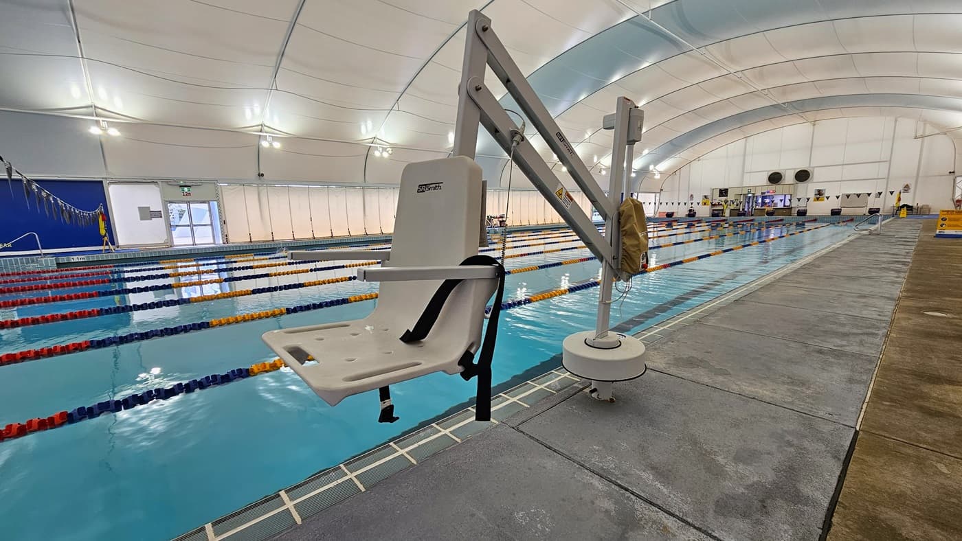 In the centre is the 8-lane lap pool, equipped with floating ropes and stairs with railings. A hoist is positioned on the pool deck, along with safety reminder banners on the right. On the far end, a glass door leads to the outdoor pool. The dome-shaped ceiling features floodlights and LED lights.