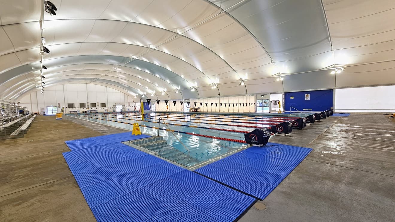 In the center is the 8-lane lap pool, equipped with floating ropes and stairs with railings. Diving boards are arranged on the pool deck. Blue matting is installed on both corners. Tiered seating is on the right. Strings of blue flags are hung across the pool. Tiered seating and safety reminder banners are visible on the pool deck area to the left. The dome-shaped ceiling features floodlights and LED lights.