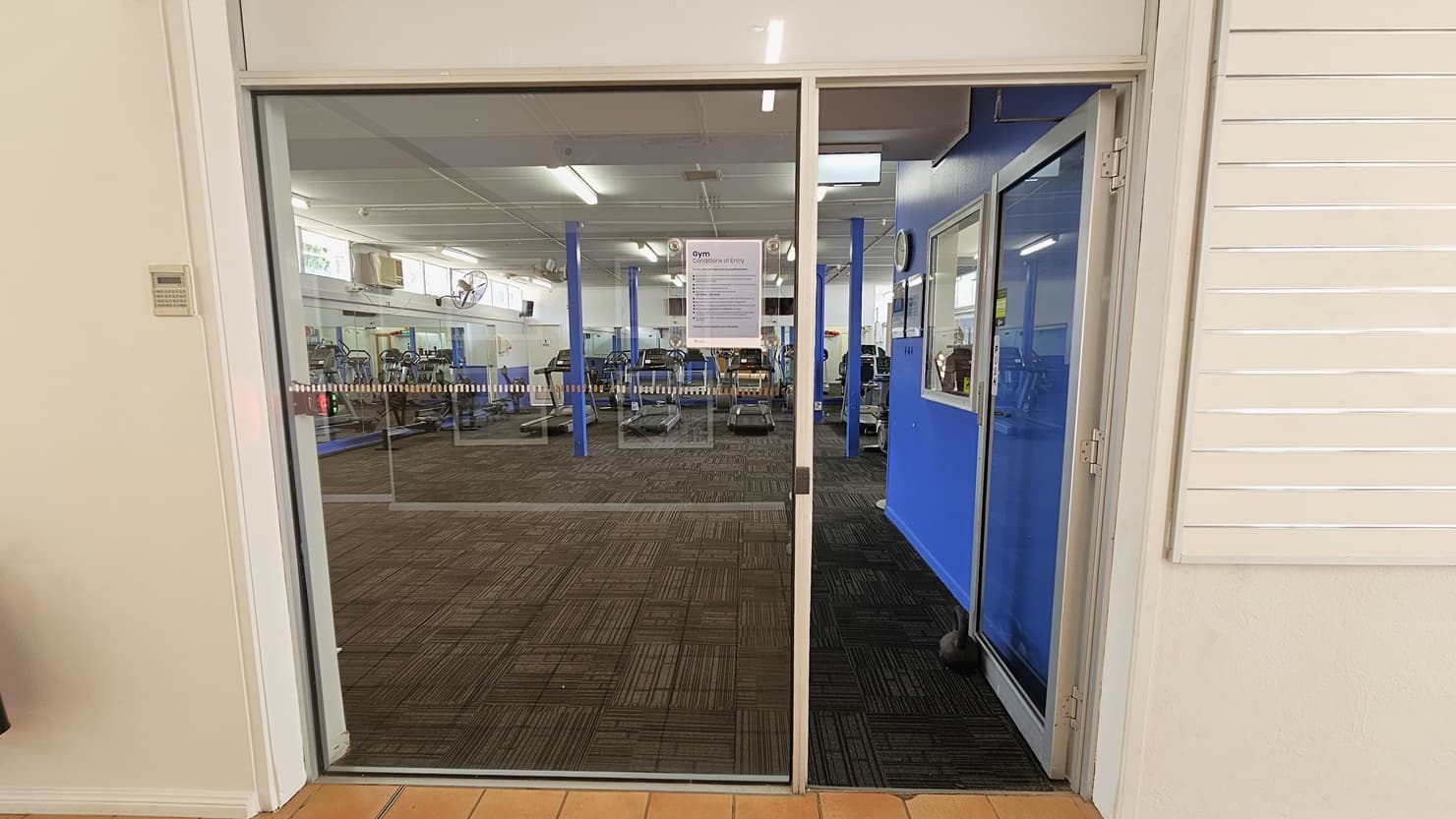 A manual glass door that opens inward leads to the gym. A poster bearing a Gym use reminder is posted on the glass wall. The external wall is all white with a linear design. Inside, the floor is covered with grey carpet. To the right is an office with a glass window and blue walls. A wall clock and framed posters are affixed to the wall. Various gym equipment is visible inside.