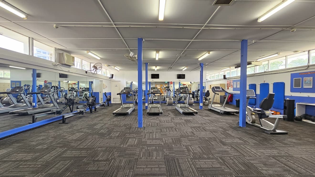 In the middle are various pieces of equipment, including treadmills and rowing machines. The left wall features floor-to-ceiling glass. To the right is a blue and white wall featuring a board with posters. The upper wall features glass windows allowing natural light and a view outside. Flat-screen TVs are mounted on the walls, along with wall fans. The ceiling is all white with fluorescent lights, and the floor has grey tiles.