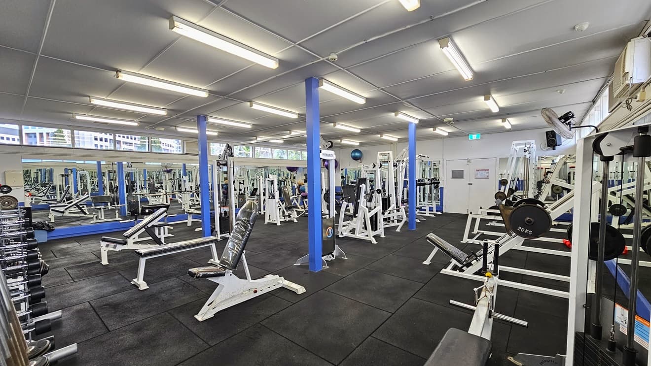 Weights are arranged on the left, and a floor-to-ceiling mirror covers the left wall. Bench chairs, Bench Press, and Smith machines are arranged in the middle and on the right side. The upper wall features glass windows allowing natural light and a view outside. Flat-screen TVs are mounted on the walls, along with wall fans. The ceiling is all white with fluorescent lights, and the floor has black rubber tiles.