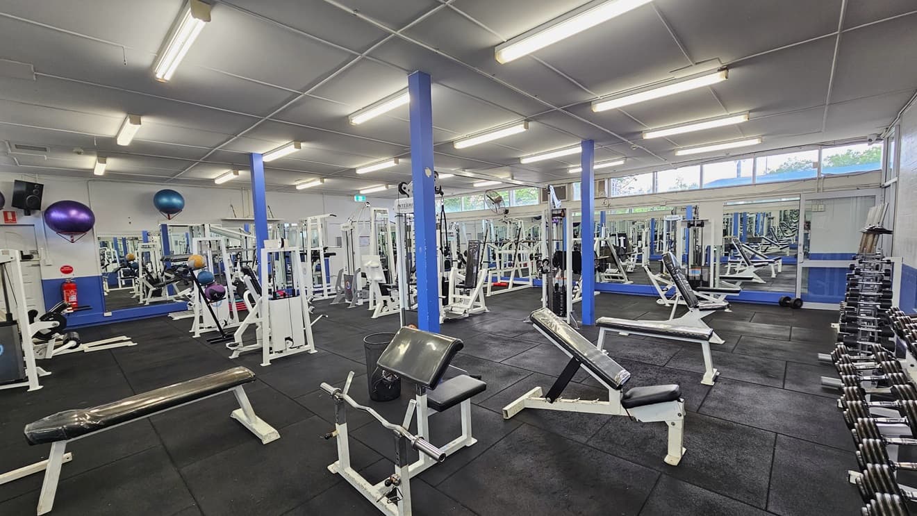 Weights are arranged on the right, and a floor-to-ceiling mirror covers the middle wall. Bench chairs, Bench Press, and Smith machines are arranged in the middle and on the right side. The upper wall features glass windows allowing natural light and a view outside. Flat-screen TVs are mounted on the left wall, along with wall fans. Stability balls are also displayed on the wall. The ceiling is all white with fluorescent lights, and the floor has black rubber tiles.