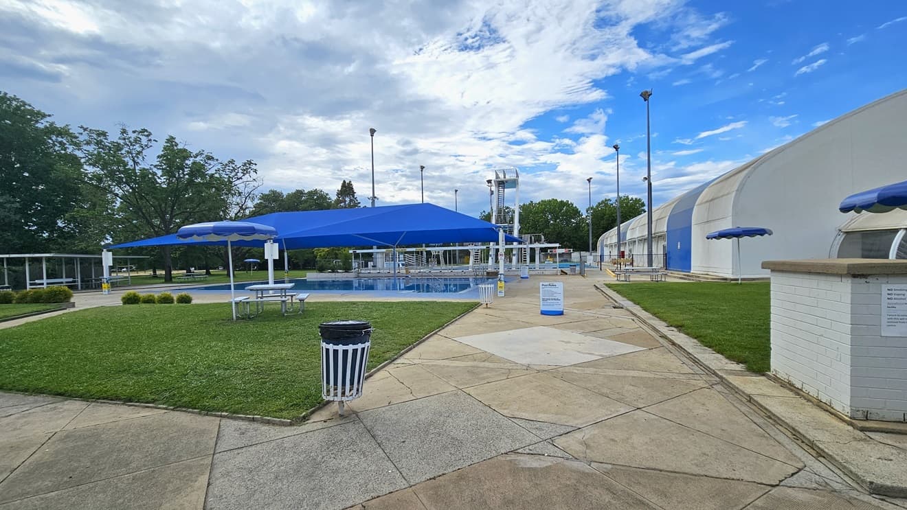 To the left, a pool covered with shade cloth. A picnic table is positioned under an umbrella shade in the manicured grass area. To the right is the dome-shaped indoor aquatic facility building. Trimmed grass also covers the right space. Further ahead, there is a shaded facility with bench chairs, and the diving pool is behind.