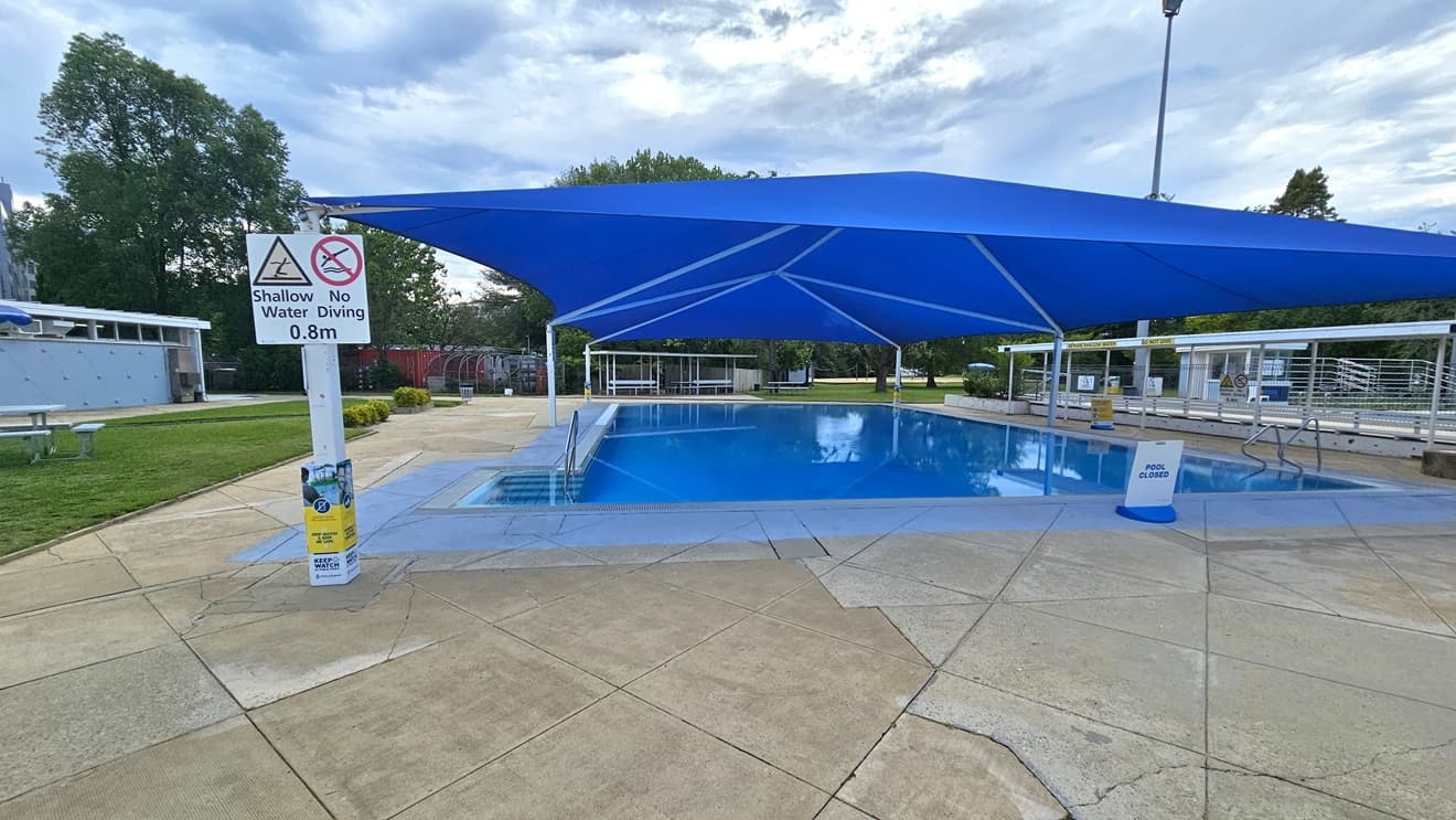 A pool is covered with a blue shade cloth. To the left is a stairs entry equipped with handrails. To the right is another handrail. Safety reminder posters are posted on the post at the left, and a free-standing banner stands in the pool deck. A blue facility building, along with a picnic table, is positioned on the left. To the far end is a shaded tiered seating. The floor is made of concrete. Trimmed grass and tall trees surround the area.
