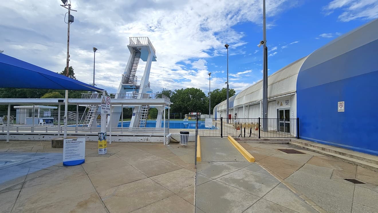 To the left is a section of the kids' pool, and to the right is a ramp leading to the diving pool. On the left is the dome-shaped blue and white structure housing the indoor aquatic center. The diving pool features four-level diving platforms. A steel gate and fence separate the diving pool from the kids' pool. A rubbish bin is positioned near the ramp. Safety banners are affixed to the wall. On the far left is a shaded facility with tiered seating. Tall outdoor lights are visible.