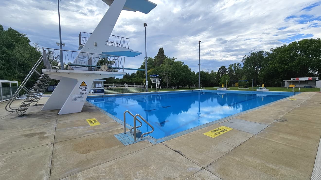The diving pool has four-level diving platforms with steel stairs and railings. A steel gate and fence separate the diving pool from the kids' pool. The pool is equipped with stairs and handrails, and a security warning denotes the water's depth. To the left is a shaded facility with tiered seating, and adjacent to it is a watchtower. To the far right is a spacious area with green grass and tall trees. Tall outdoor lights are visible.