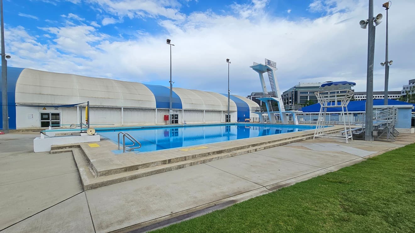 The pool is equipped with stairs and handrails. To the right is a watchtower, and next to it is tiered seating. To the left is the blue-white dome-shaped building housing the indoor aquatic facilities. On the left is a shaded facility with tiered seating, and adjacent to it is a watchtower. The diving platform is located in the middle, equipped with steel stairs and railings. Neatly trimmed grass surrounds the right side of the pool.