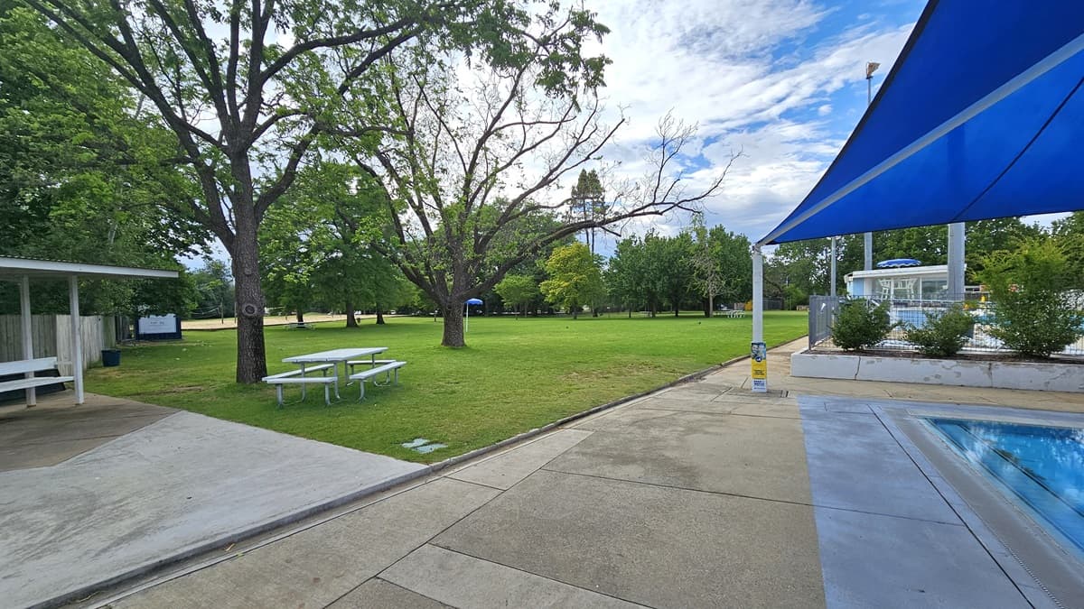 To the left is a shaded facility with picnic tables underneath. More picnic tables are positioned under the shade of trees. Trimmed grass and tall trees cover the left section. To the right is the kids' pool covered with a blue shade cloth. Adjacent is a metal fence that separates the diving pool.