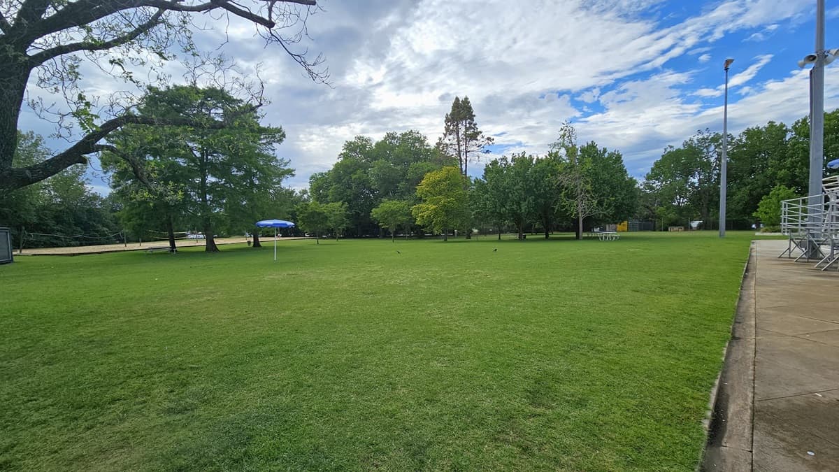 To the left is a spacious area covered with green grass and tall trees. Picnic tables are placed under the trees. An umbrella shade is visible. To the right is tiered seating and the watchtower.