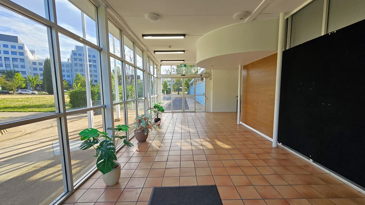 The doorway features a black anti-slip mat. The left side is glass, offering an exterior view. Potted ornamental plants line up along the wall. The right wall combines brown concrete and brown tiles. At the corridor's end is another glass door. The ceiling is uniformly white with fluorescent and LED lighting, and the floor is covered in brown tiles.