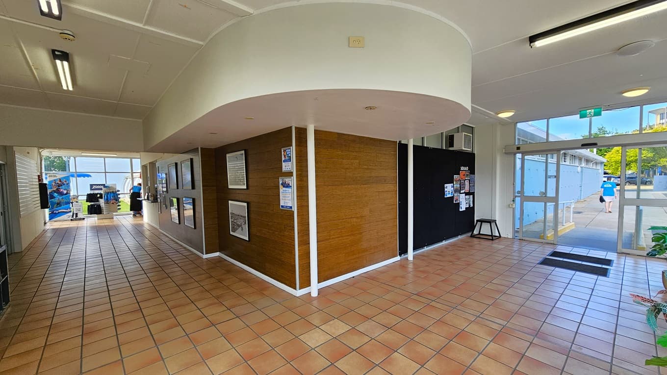 The L-shaped corridor features brown tiles. On the right is the entrance with a glass door and non-slip mats. A blackboard displays posters, and ornamental plants are arranged on the right. The middle wall, with a timber design, showcases large framed posters. At the corridor's end are displayed retail items. The ceiling is white with fluorescent lights.