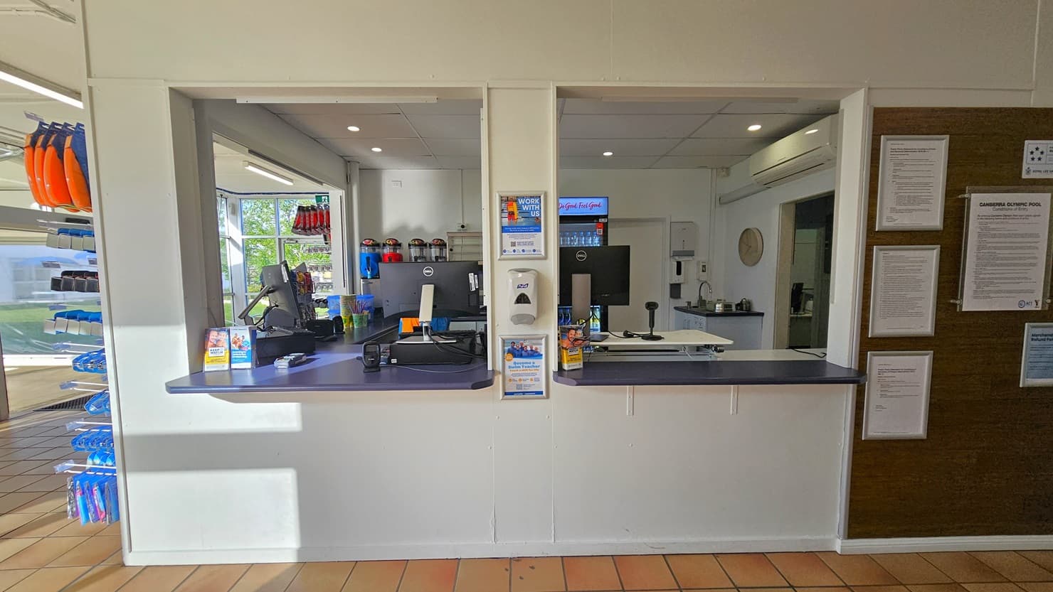 The reception features three large windows. The desk is equipped with computers and swiping machines, with a fridge displaying cold drinks behind. On the right, a brown board displays framed posters, and on the left, a rack showcases goggles and other swimming accessories. The walls and ceiling are white, and the floor has brown tiles.