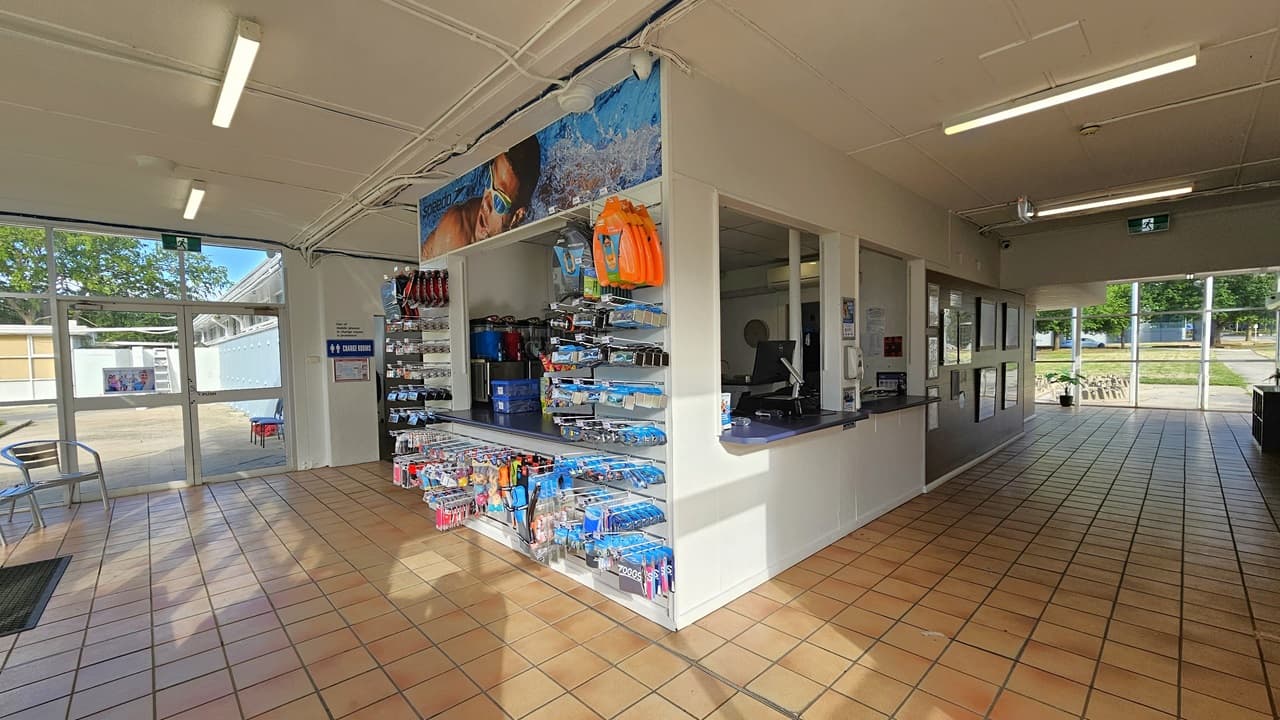 The reception has three large windows. The desk is equipped with computers, swiping machines, and a fridge displaying cold drinks. On the right, a brown board displays framed posters, while the left features a rack showcasing goggles and swimming accessories. Movable chairs and an anti-slip mat are positioned on the right. The walls and ceiling are white, and the floor has brown tiles.
