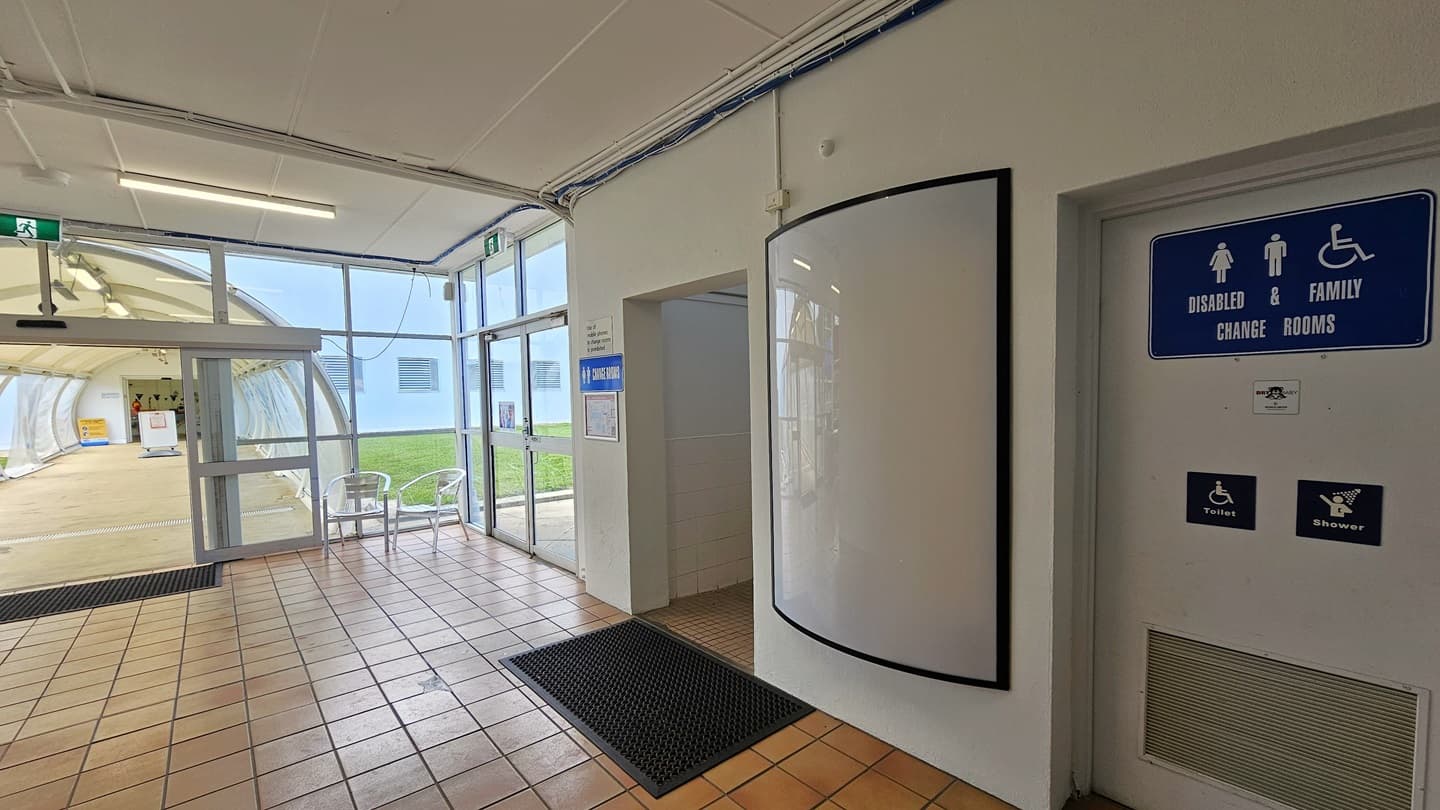 To the right are accessible bathrooms with signage and black rubber mats on the doorsteps. To the left, an automatic glass door leads to a bridge with a dome-shaped roof, featuring two movable chairs in the corner. Another glass door is to the right. The floor has brown tiles, and the ceiling and walls are white.