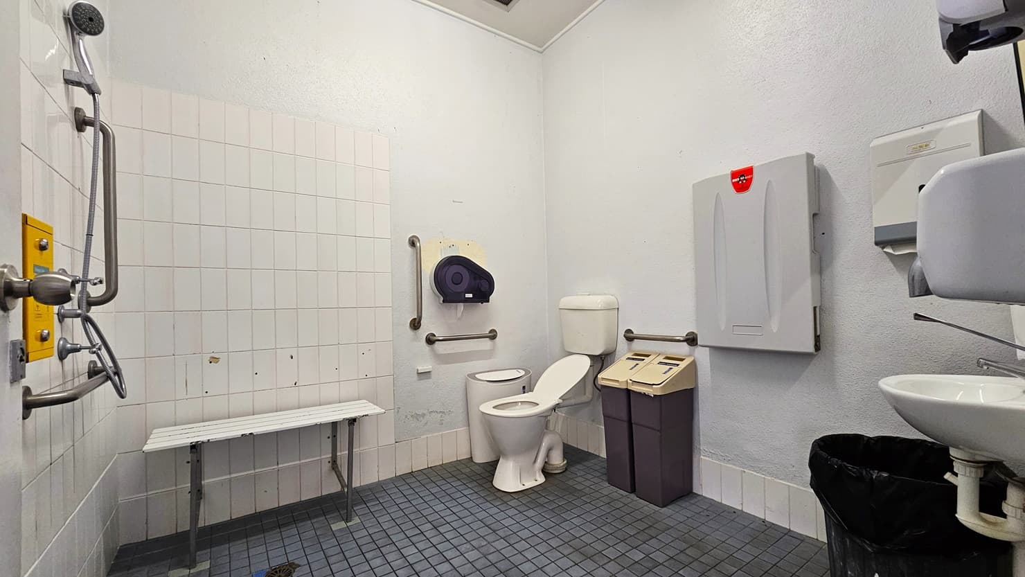 Inside the accessible bathroom, the toilet bowl equipped with handrails is positioned in the corner, accompanied by two rubbish bins. A foldable change table and a tissue dispenser are mounted on the wall. To the left, there is a shower, and adjacent to it is a bench chair. To the right, a wash basin, a liquid soap dispenser, a hand dryer, and a tissue dispenser are mounted on the wall. The walls and ceiling are all white, and the floor has grey tiles.