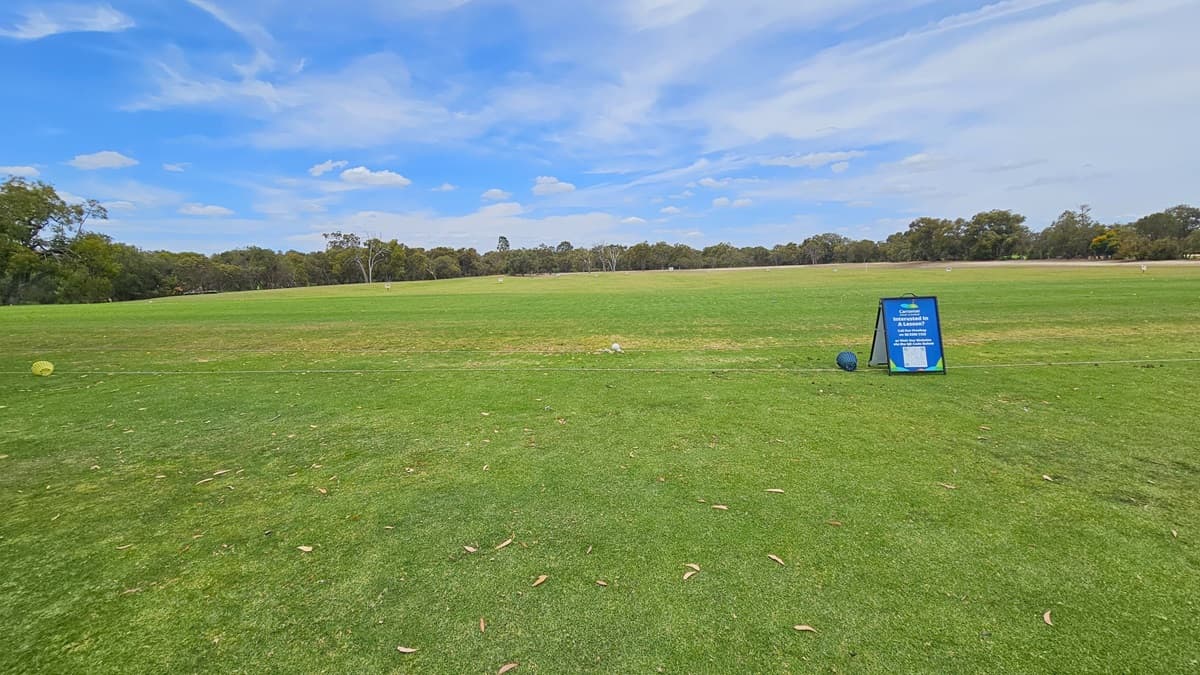 A concrete path directs towards the green. On the right, there is an elevated grassy area with a green sign reading "10th Tee." A golfer is visible near the freestanding banner. On the left, there is manicured grass. Further ahead, more trees are present.