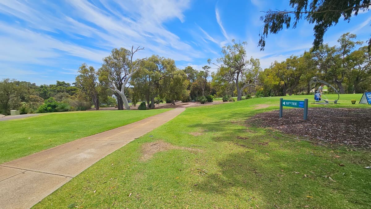 A concrete path directs towards the green. On the right, there is an elevated grassy area with a green sign reading "10th Tee." A golfer is visible near the freestanding banner. On the left, there is manicured grass. Further ahead, more trees are present.