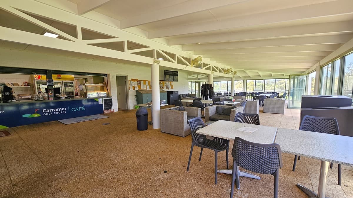 On the left, the cafe counter exhibits the facility's signage with a food rack. Adjacent is a corkboard for reminders, and cleaning accessories are underneath. To the right, there are cane-style couches and movable chairs and tables. A black rubbish bin is positioned near the middle post. The wall is made of white bricks, the ceiling is white, and the floor has brown tiles, featuring a large floor mat with the facility's name.