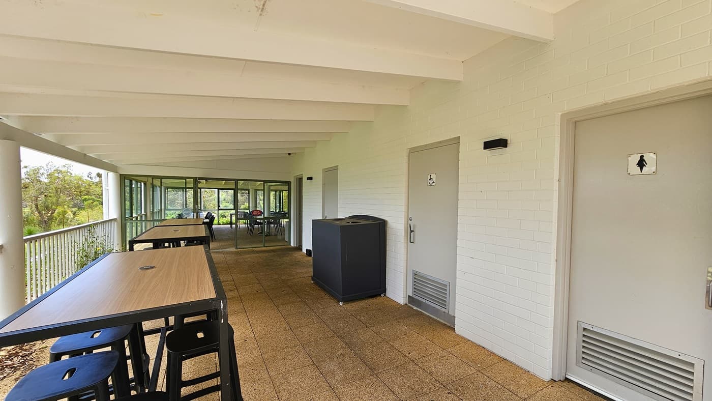 Movable chairs and tables are arranged on the left, secured by a long metal railing. To the right, three grey doors each feature gender access signage. A black rubbish bin is positioned against the white brick wall. The ceiling is white with exposed wooden beams, and the floor is covered with brown tiles. On the far end, a glass door leads to additional seating.