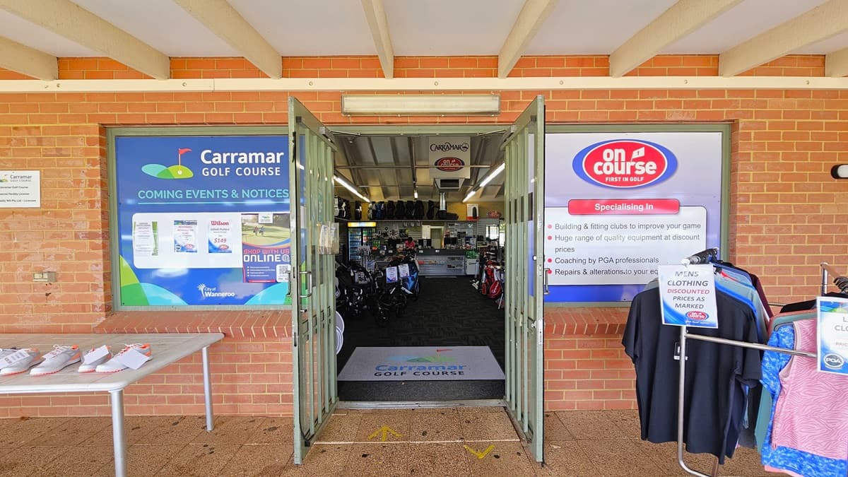 The building's facade is constructed with red bricks, adorned with banners on both sides. On the left, a white table showcases white golf shoes for sale. To the right, men's and ladies' clothing is on display. Inside, the concrete brick exterior transitions to carpeting. A large floor mat with the facility's name is at the doorstep. Retail items are on display inside.