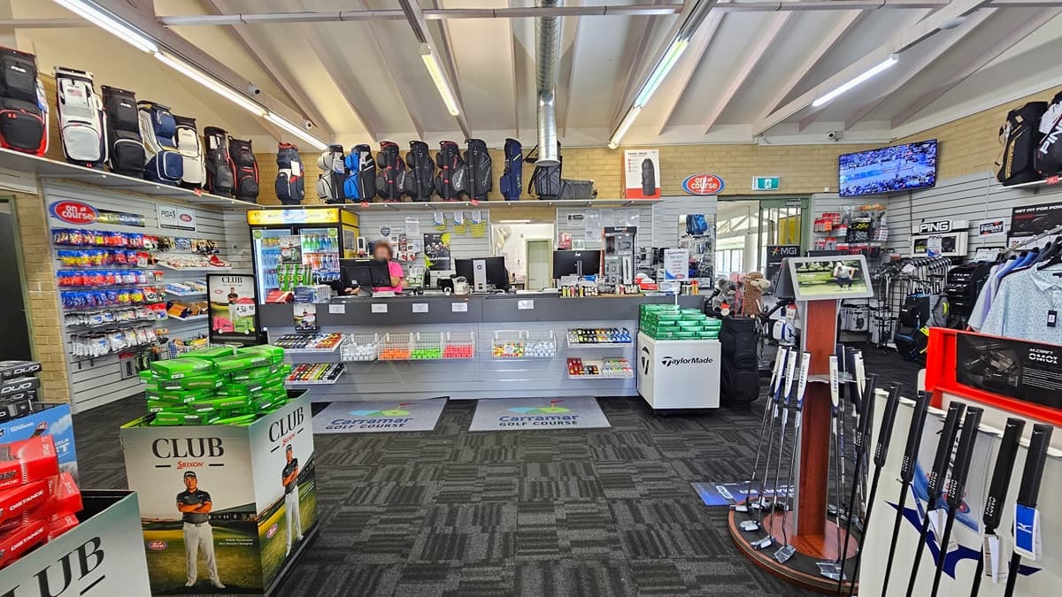 The reception counter, positioned centrally, features three computers and retail items on the desk. A fridge with cold drinks is situated behind the counter. In the upper right corner, a flat-screen TV is mounted. Golf balls and bags are displayed on the left, while golf clubs and polo shirts are arranged on the right, along with additional bags on a display rack. The ceiling is white with exposed metal beams and fluorescent lights, and the floor is covered with grey carpet.