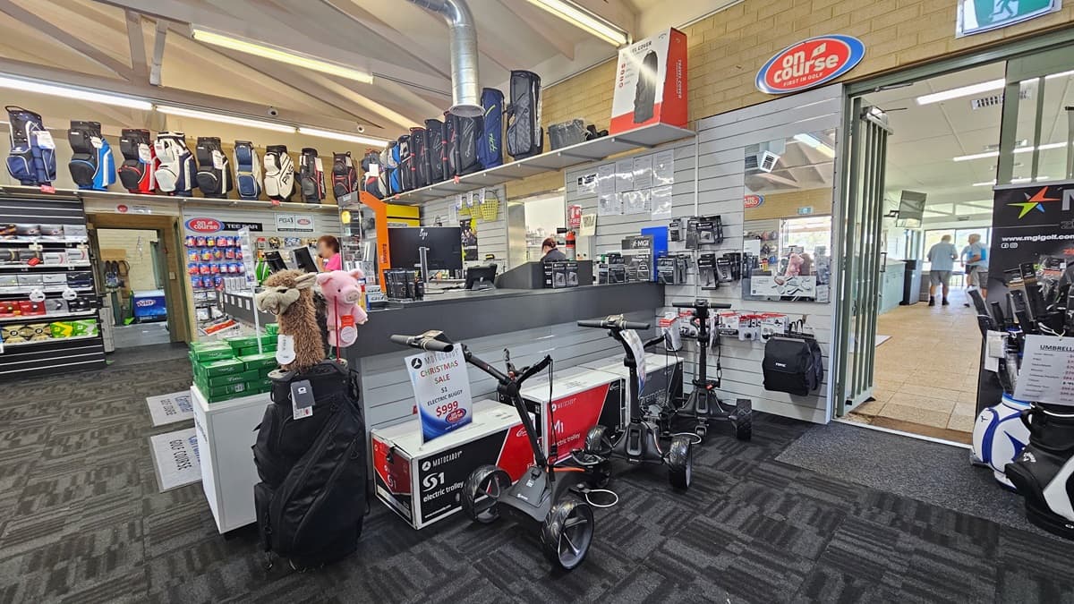 The reception counter, centrally positioned, accommodates three computers and retail items. Golf balls and bags are displayed on the left, while electric trolleys are showcased on the right. A glass door on the right opens outward, leading to an area with cream tiles. The ceiling is white with exposed metal beams and fluorescent lights, and the floor is covered with grey carpet.