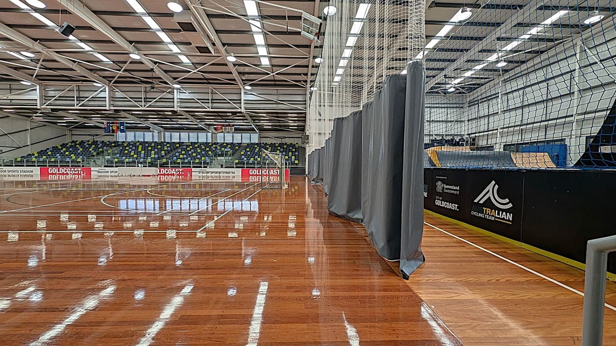 A large indoor sports hall with a polished wooden floor. On the left, there's a white wall with a fire exit door and a green "Push bar to open" sign. Above the door is an emergency exit light. A fire hose reel is mounted on the wall. In the middle, there are tiered seating with multicoloured chairs. The ceiling is high with exposed beams and fluorescent lights. On the right, a grey curtain partially covers the view of the court.