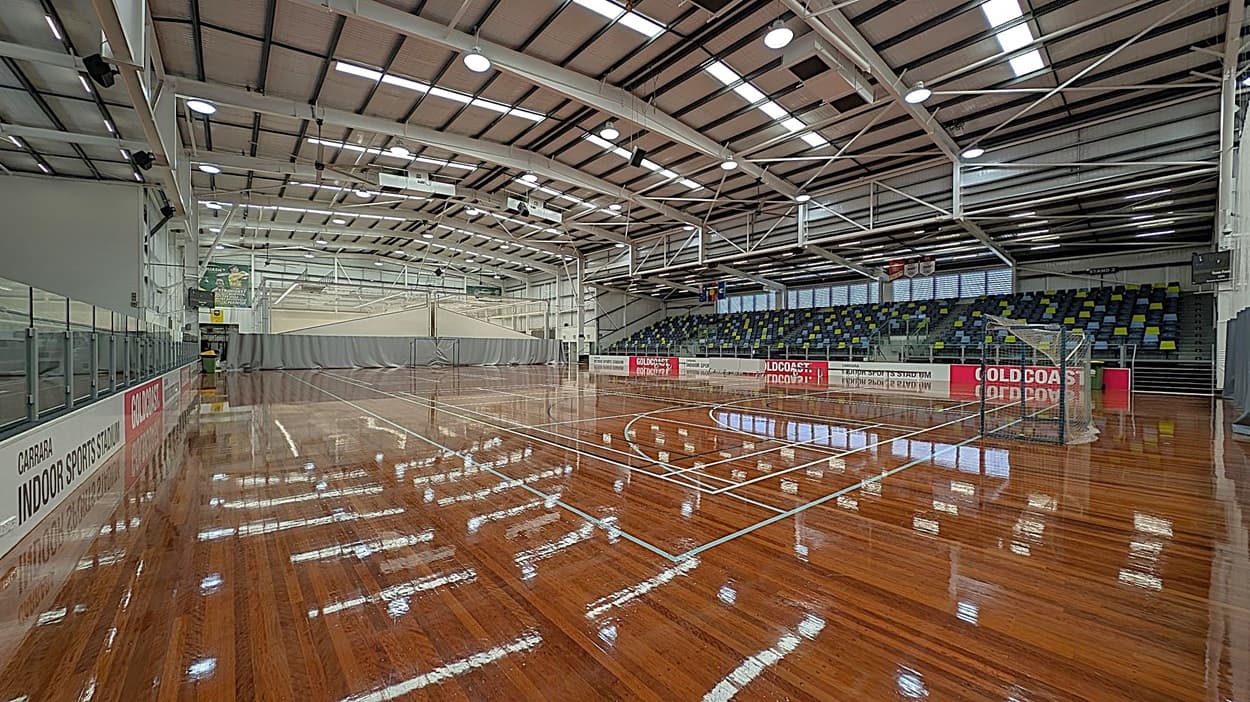 A spacious indoor sports stadium with a polished wooden floor reflecting overhead lights. On the right, there are tiered seating areas with multicoloured seats. A netted goal is positioned near the seating. On the left, there are barriers with advertising banners. The ceiling is high with exposed beams and numerous lights. In the background, a large curtain divides the space, and there are additional sports equipment and banners visible.