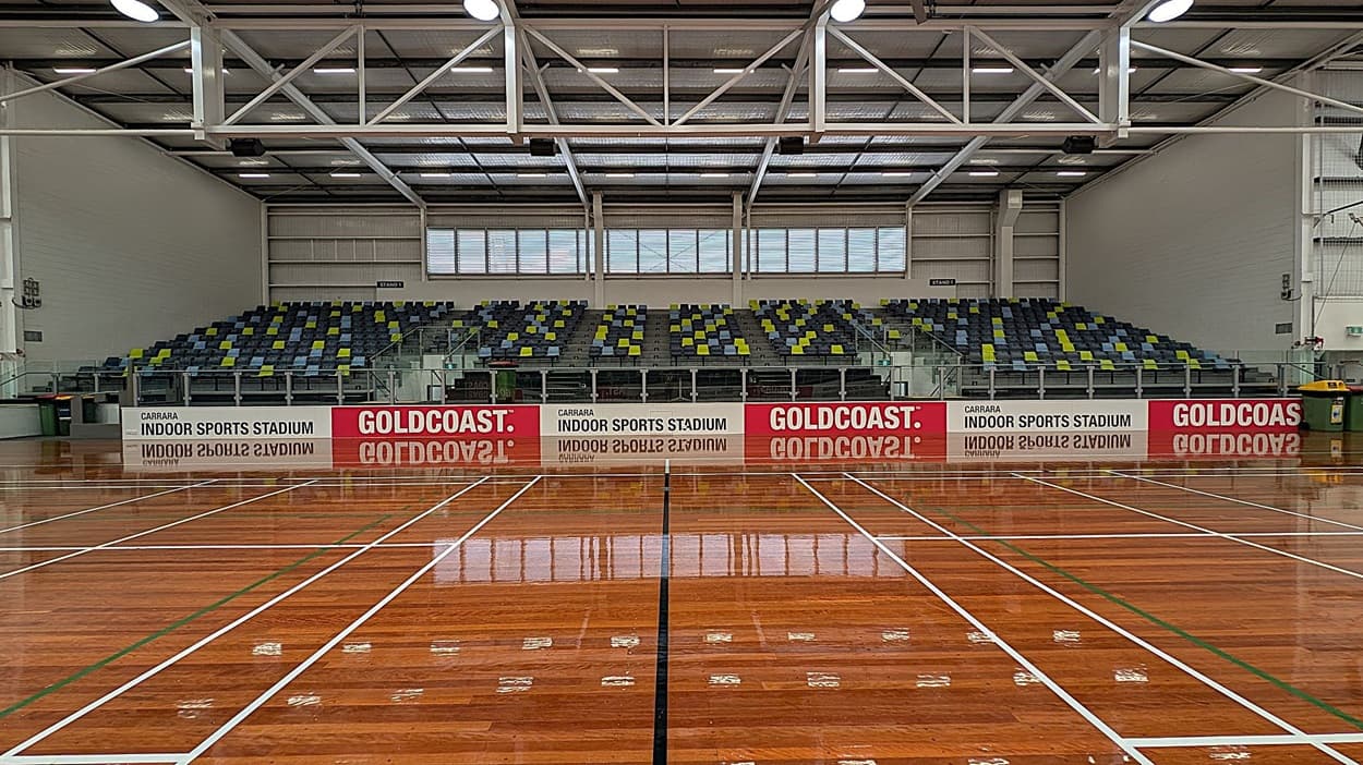 A spacious indoor sports stadium with a polished wooden floor marked with white and green lines. In the background, there are tiered seating areas with multicoloured seats in shades of grey, green, and blue. Above the seats, large windows allow natural light to enter. The ceiling features exposed beams and lighting fixtures. Signs on the barrier in front of the seating read "Carrara Indoor Sports Stadium" and "Gold Coast."