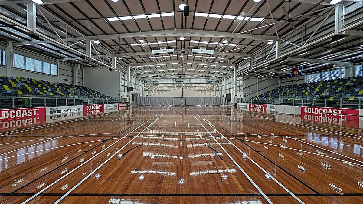 A spacious indoor sports stadium with a polished wooden floor marked for various sports. On the left and right are tiered seating areas with green, blue, and yellow seats. The walls have large windows and banners reading "GOLDCOAST" and "INDOOR SPORTS STADIUM." The ceiling is high with exposed beams and lighting fixtures. In the centre, a grey curtain divides the space.