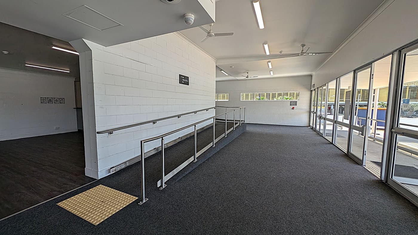 A corridor with a ramp on the left, featuring a metal handrail and a yellow tactile paving at the base. The floor is carpeted in dark grey. On the right, there are large glass doors and windows allowing natural light to enter. The ceiling is white with fluorescent lights and ceiling fans. The walls are painted white, and there is a small sign on the left wall. In the background, there are more windows and a view of an outdoor area.