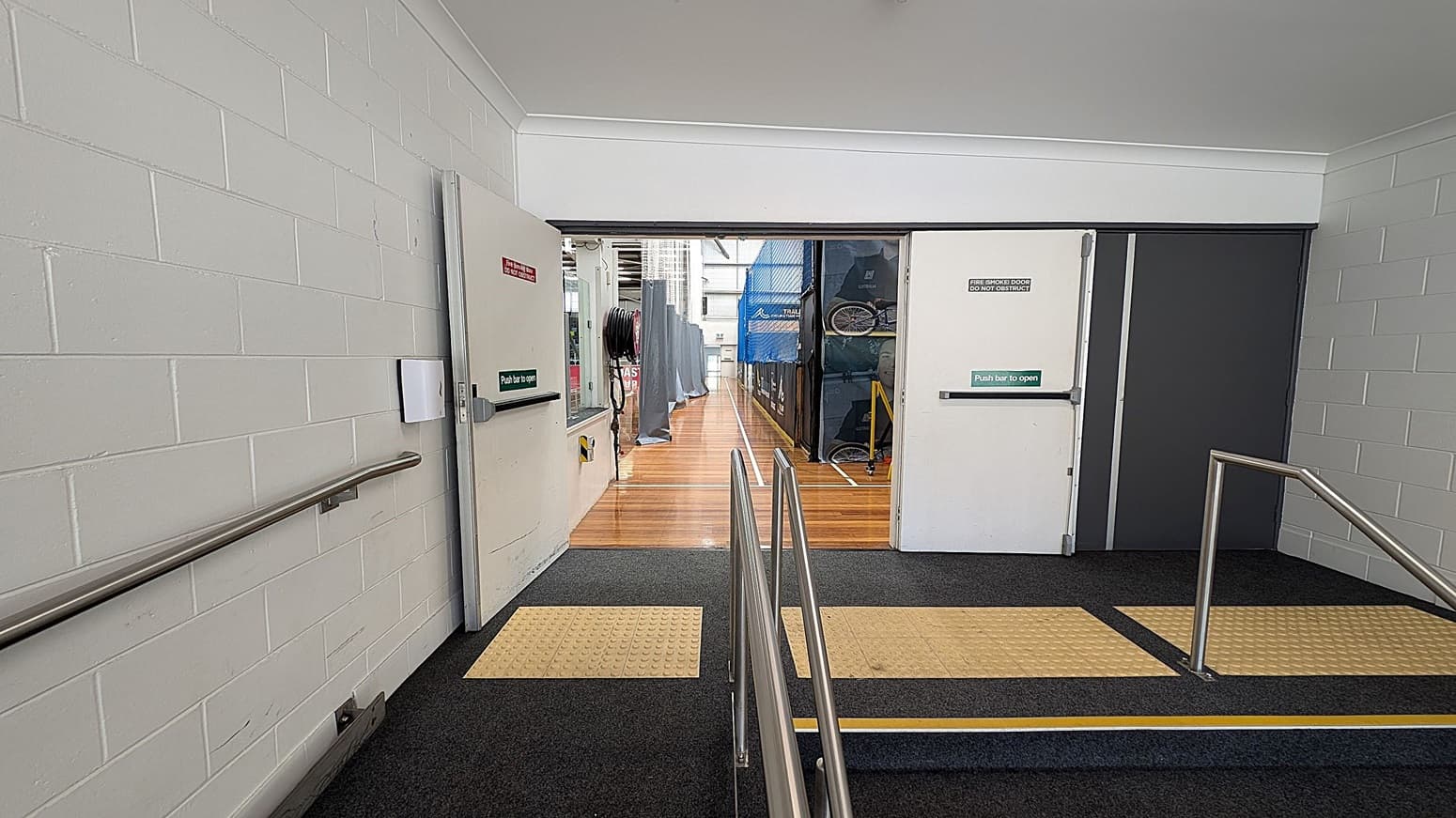 A corridor with white brick walls and a grey carpeted floor. On the left, there's a metal handrail along the wall. In the centre, two open doors with "Push bar to open" signs lead to a wooden-floored area with sports equipment, including a blue net and bicycles. The right side also has a metal handrail. The ceiling is plain white.