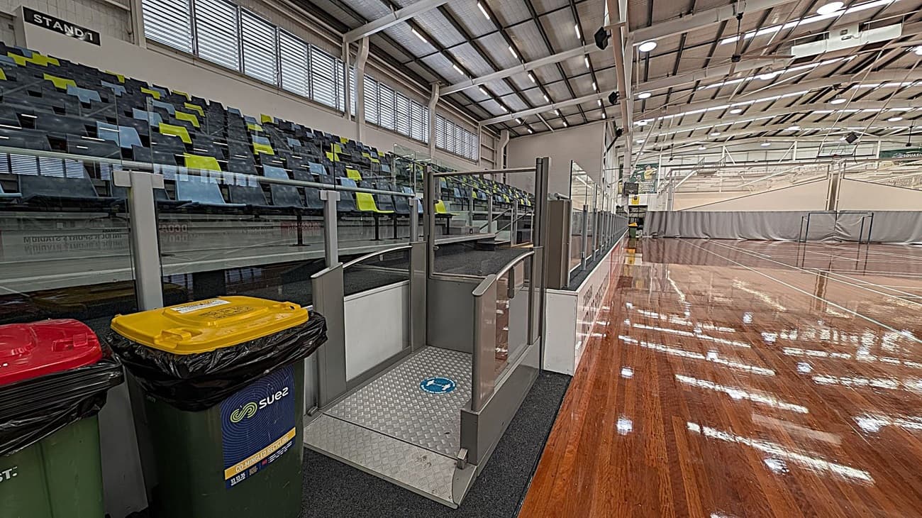 Indoor sports hall with a polished wooden floor on the right. On the left, there are tiered seating with alternating blue and green seats. In the foreground, there are three bins with red, yellow, and green lids. A ramp with metal railings leads up to the seating area. The ceiling is high with exposed beams and multiple lights. Large windows with horizontal slats are above the seating area.