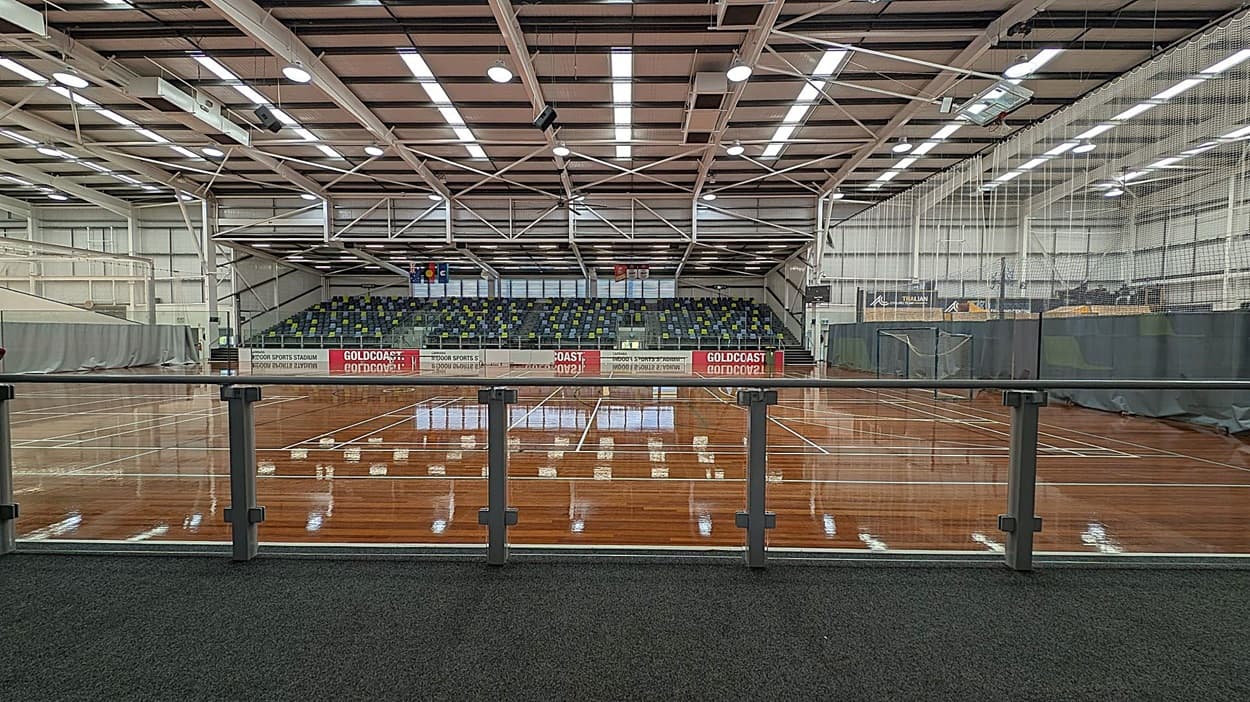 A large indoor sports stadium with a polished wooden floor and multiple marked courts. In the background, there are tiered seating areas with multicoloured seats. The ceiling is high with exposed beams and bright overhead lighting. A glass railing runs along the foreground, separating the viewing area from the courts. Banners and nets are visible on the right side, and the walls are lined with grey curtains.